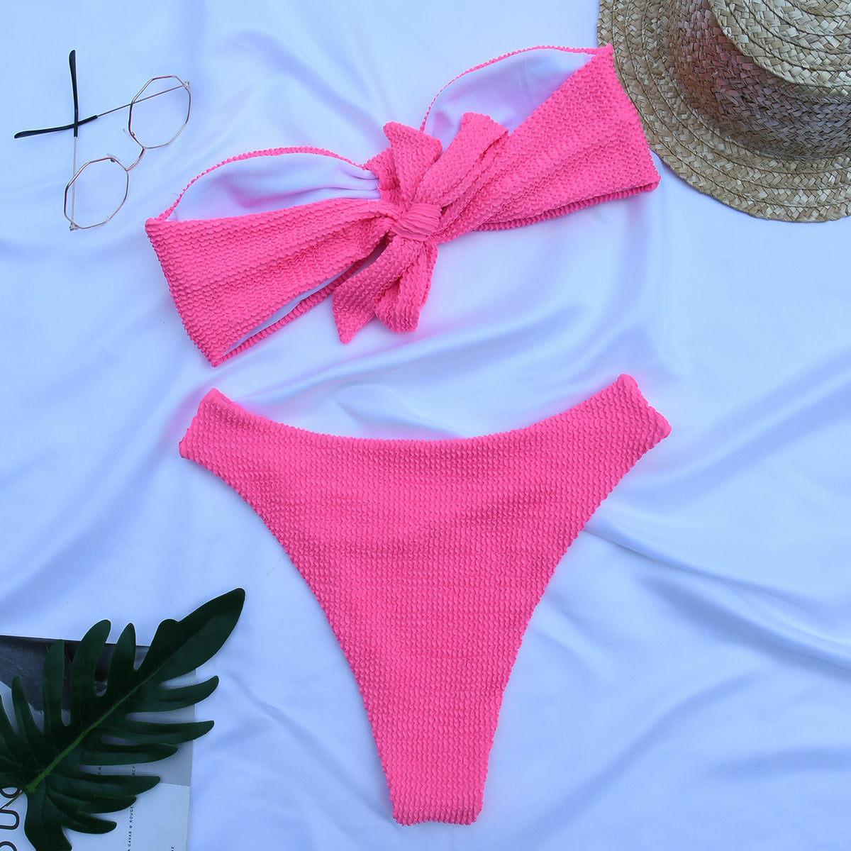 Pink textured bandeau bikini set with large front bow detail laid flat with sunglasses and straw hat
