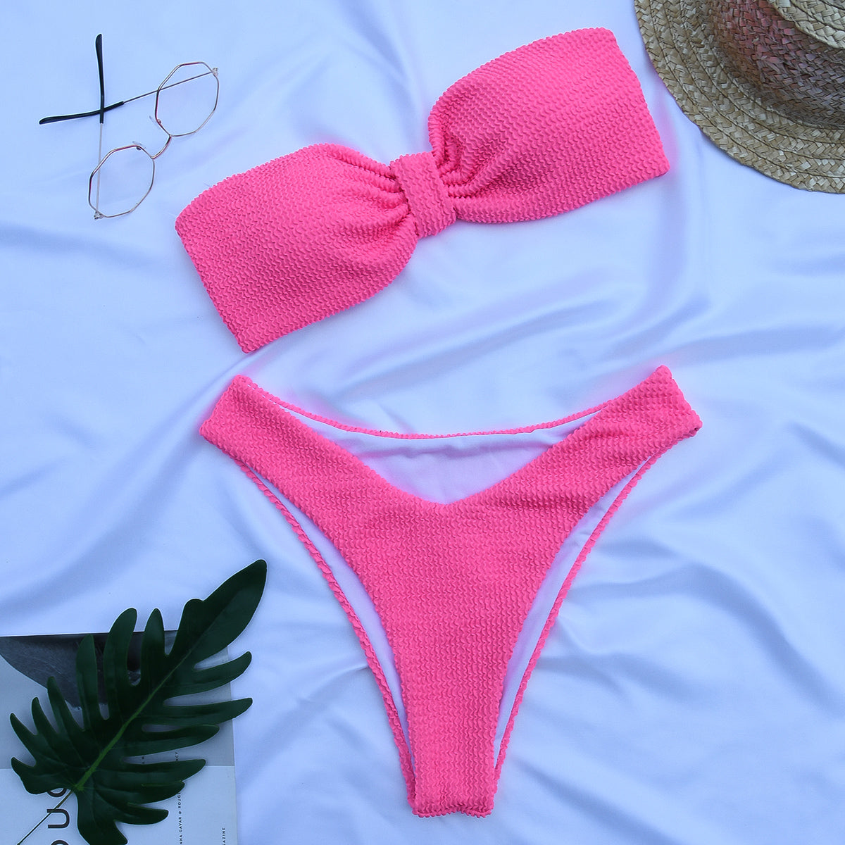 Pink textured bandeau bikini set laid flat with sunglasses and straw hat on white fabric